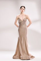 Photograph of a slender, fair-skinned woman with dark brown hair styled in a low bun. She wears a floor-length, champagne-colored, sequin-embellished gown with spaghetti straps and a deep V-neckline, accentuating her modest bust. The dress has a fitted bodice and a flowing, slightly shimmering skirt. She stands against a simple, white background with soft lighting, creating a minimalistic and elegant atmosphere. Her hands are placed on her hips, and she has a poised, confident expression.