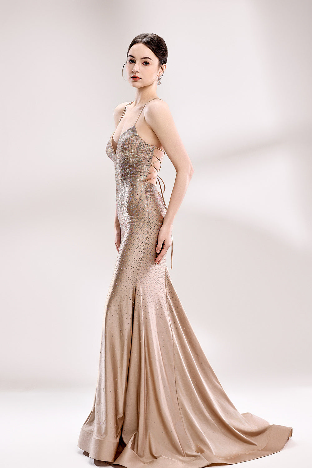Photograph of a slender, fair-skinned woman with dark brown hair styled in a low bun. She wears a floor-length, champagne-colored, sequin-embellished gown with spaghetti straps and a deep V-neckline, accentuating her modest bust. The dress has a fitted bodice and a flowing, slightly shimmering skirt. She stands against a simple, white background with soft lighting, creating a minimalistic and elegant atmosphere. 