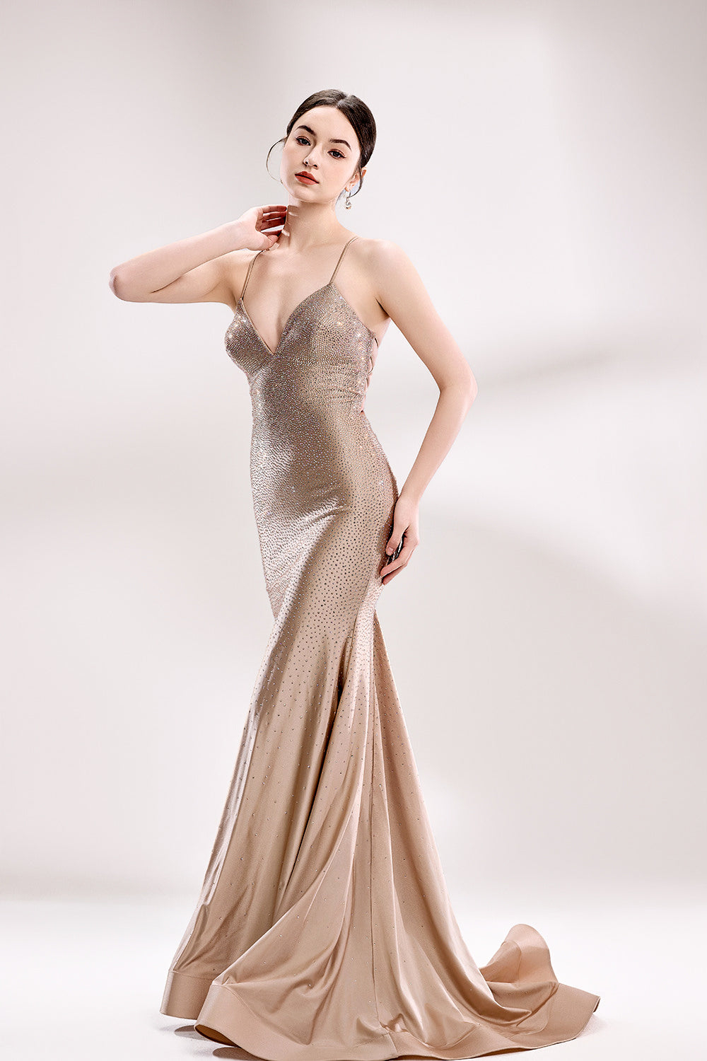 Photograph of a slender, fair-skinned woman with dark hair styled in a sleek updo, wearing a shimmering, metallic bronze, floor-length gown with a plunging V-neckline and thin straps. She poses elegantly against a plain white background, with one hand resting on her neck and the other on her hip. The gown's fabric has a subtle sparkle, catching the light. She wears large, round hoop earrings, adding a touch of sophistication. The overall style is glamorous and sophisticated, highlighting the elegance of the