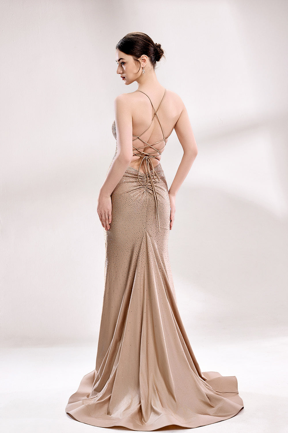 Photograph of a slender woman with fair skin and dark hair styled in a sleek bun, wearing a floor-length, metallic taupe gown with crisscross straps and a high slit. The gown features intricate beadwork and a delicate lace-up detail at the back. She stands against a minimalist, white background, with soft, diffused lighting highlighting the gown's shimmering texture. Her pose is elegant, with one hand resting on her hip and the other hanging loosely by her side.