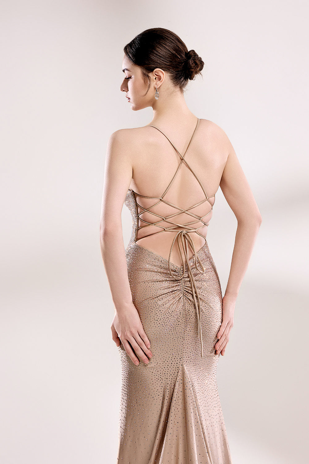Photograph of a slender woman with fair skin and dark hair styled in a sleek bun, wearing a floor-length, metallic taupe gown with crisscross straps and a high slit. The gown features intricate beadwork and a delicate lace-up detail at the back. She stands against a minimalist, white background, with soft, diffused lighting highlighting the gown's shimmering texture. Her pose is elegant, with one hand resting on her hip and the other hanging loosely by her side.