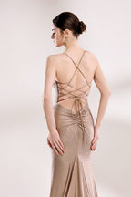 Photograph of a slender woman with fair skin and dark hair styled in a sleek bun, wearing a floor-length, metallic taupe gown with crisscross straps and a high slit. The gown features intricate beadwork and a delicate lace-up detail at the back. She stands against a minimalist, white background, with soft, diffused lighting highlighting the gown's shimmering texture. Her pose is elegant, with one hand resting on her hip and the other hanging loosely by her side.