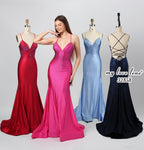 This photograph features three mannequins showcasing elegant evening gowns against a plain white background. The central mannequin wears a bright pink gown with intricate beadwork, while the left one dons a deep red gown with a similar design. The right mannequin is adorned in a royal blue gown with a crisscross lace back. A woman stands between the mannequins, wearing the pink gown, smiling confidently. The dresses are sequined and form-fitting, emphasizing their glamorous appeal.