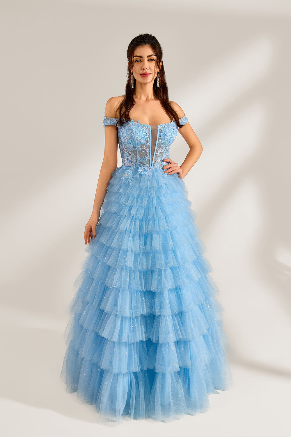 Photograph of a young woman with light skin and dark brown hair styled in loose waves. She wears a voluminous, light blue, multi-layered tulle gown with off-the-shoulder sleeves, intricate lace detailing on the bodice, and a fitted waist. She stands against a plain white background with soft shadows.1