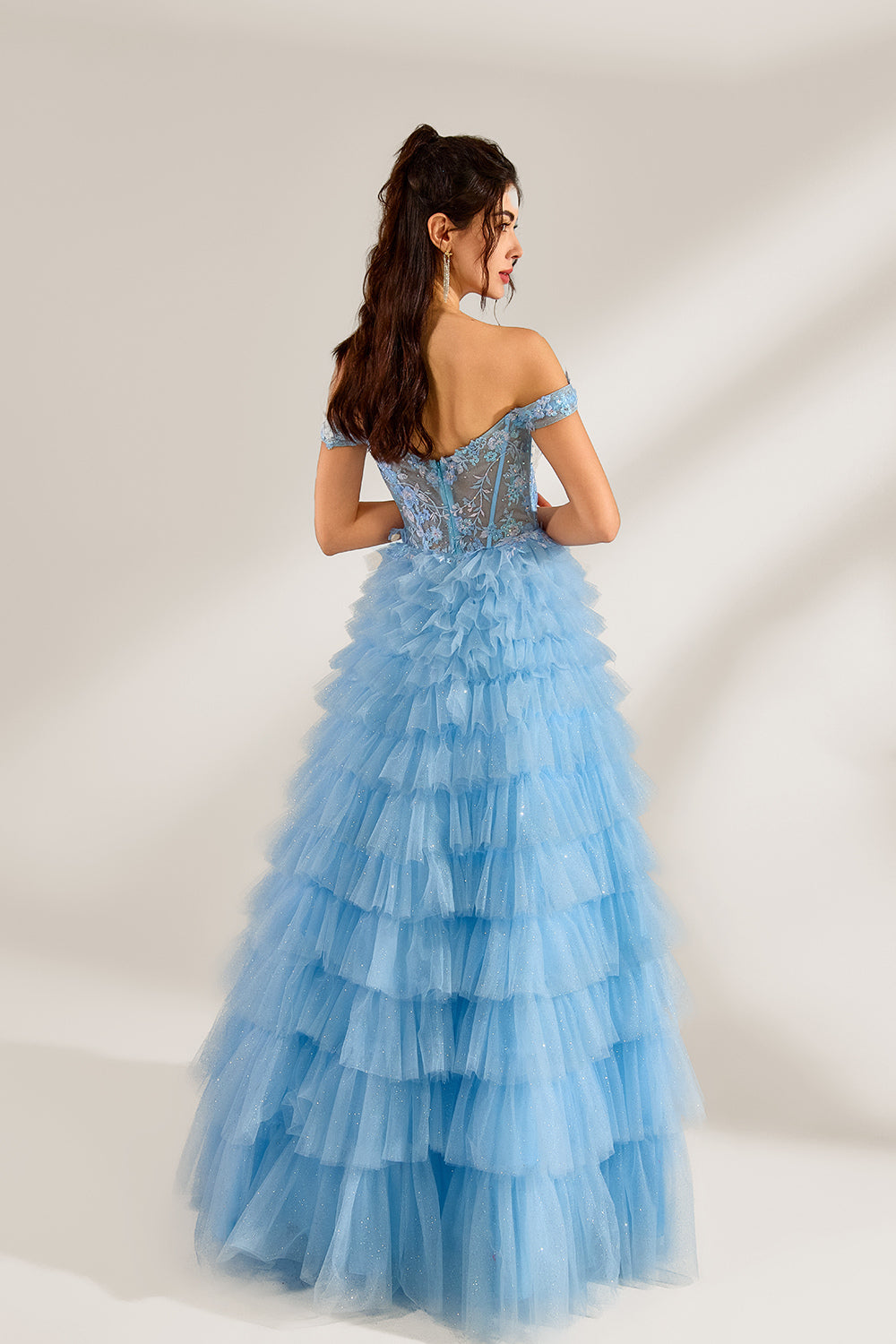 Photograph of a young woman with light skin and dark brown hair styled in loose waves. She wears a voluminous, light blue, multi-layered tulle gown with off-the-shoulder sleeves, intricate lace detailing on the bodice, and a fitted waist. She stands against a plain white background with soft shadows.2