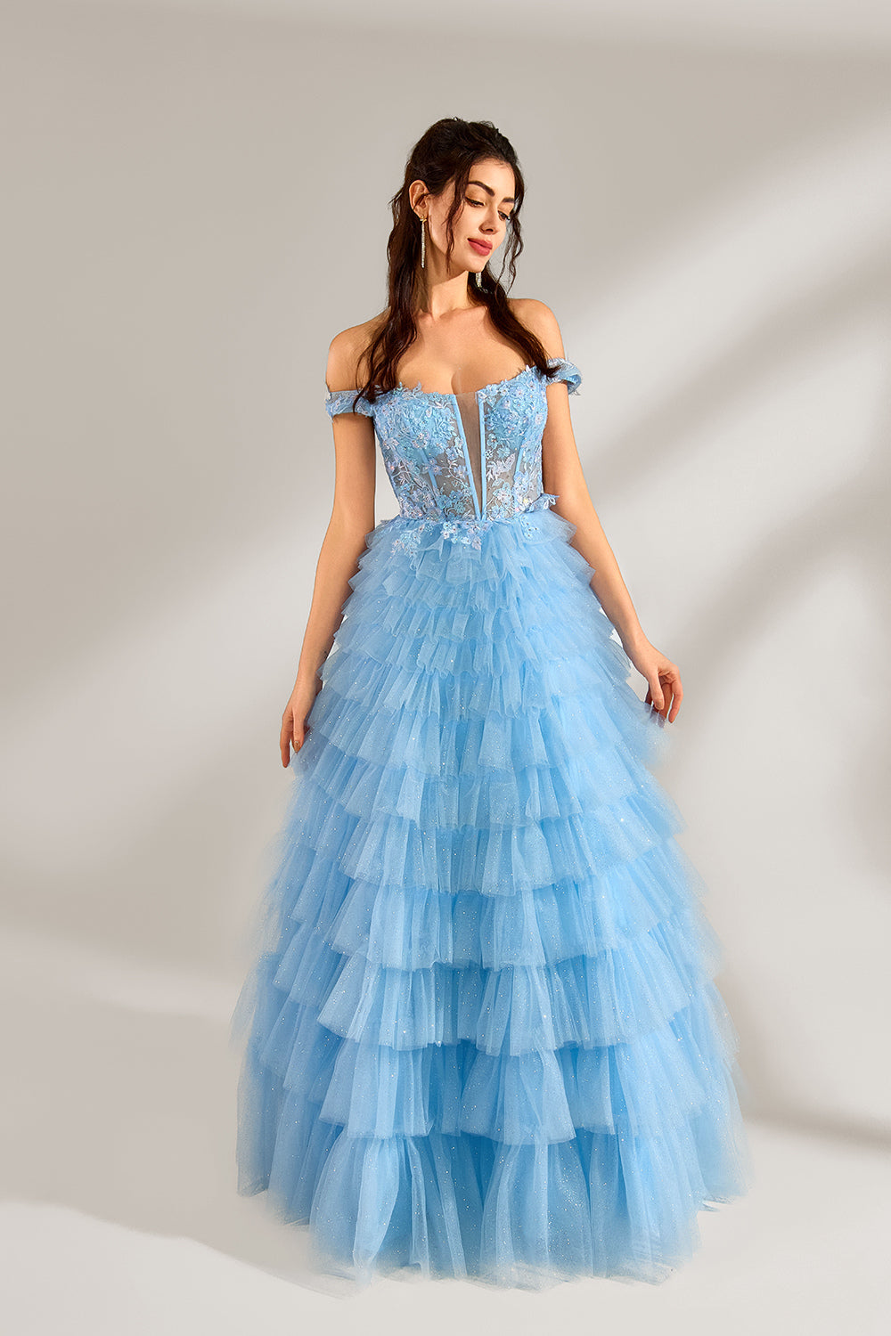 Photograph of a young woman with light skin and dark brown hair styled in loose waves. She wears a voluminous, light blue, multi-layered tulle gown with off-the-shoulder sleeves, intricate lace detailing on the bodice, and a fitted waist. She stands against a plain white background with soft shadows.3