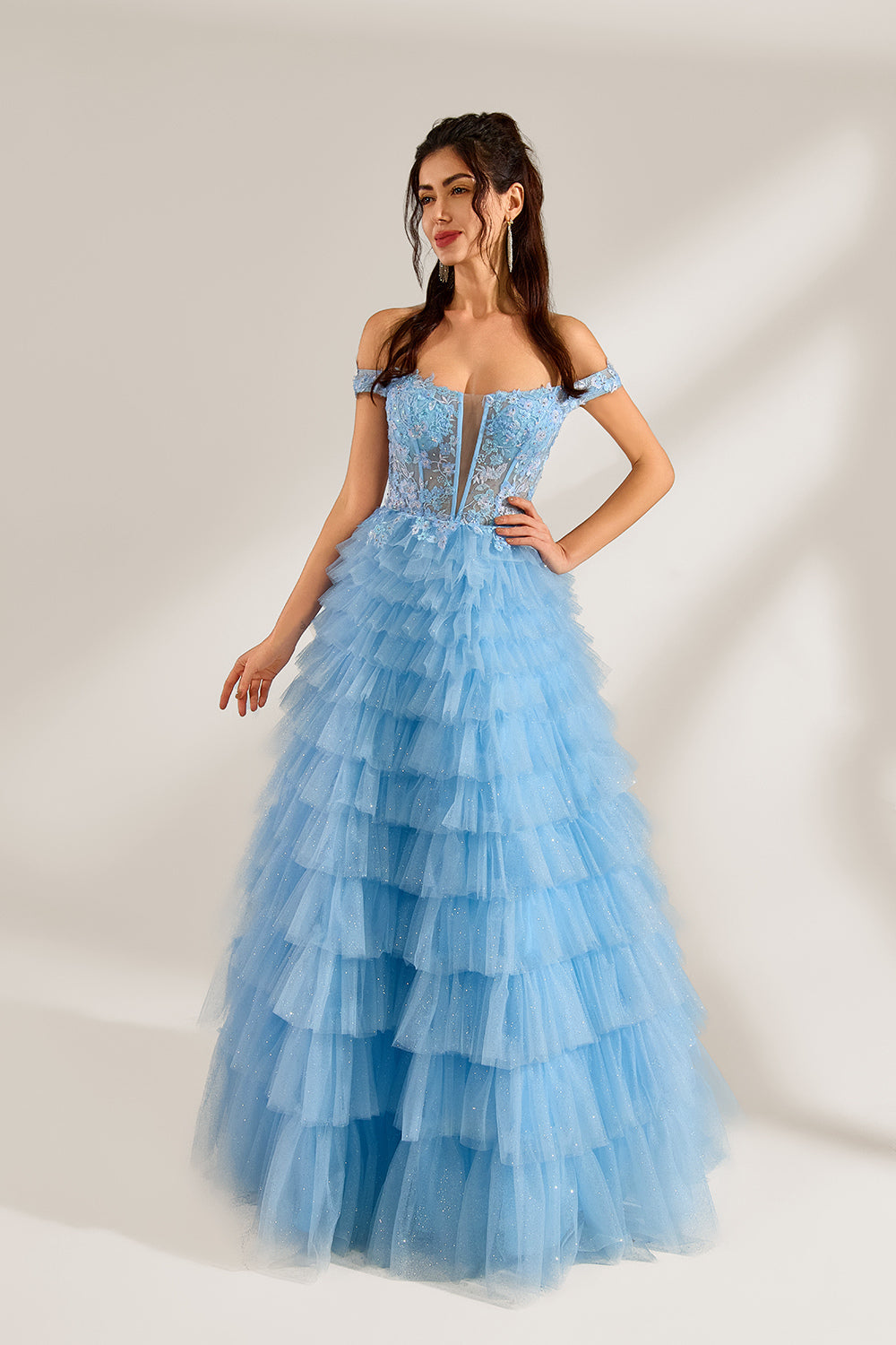 Photograph of a young woman with light skin and dark brown hair styled in loose waves. She wears a voluminous, light blue, multi-layered tulle gown with off-the-shoulder sleeves, intricate lace detailing on the bodice, and a fitted waist. She stands against a plain white background with soft shadows.4