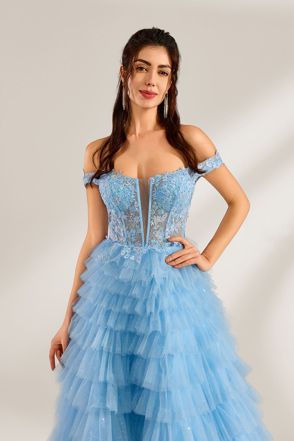 Photograph of a young woman with light skin and dark brown hair styled in loose waves. She wears a voluminous, light blue, multi-layered tulle gown with off-the-shoulder sleeves, intricate lace detailing on the bodice, and a fitted waist. She stands against a plain white background with soft shadows.5