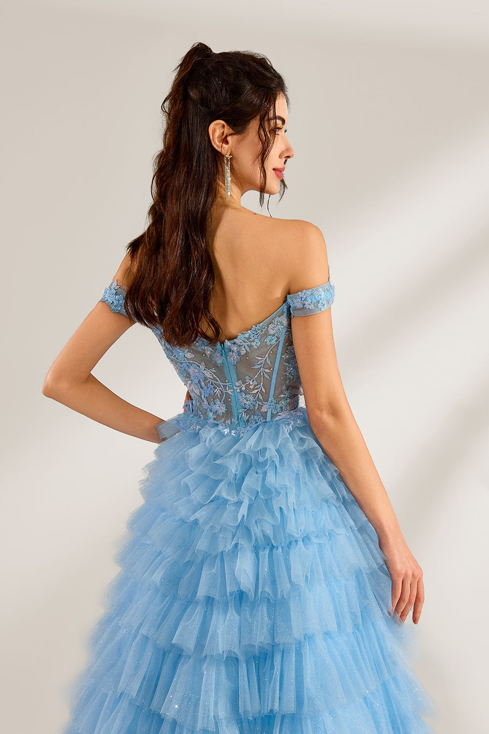 Photograph of a young woman with light skin and dark brown hair styled in loose waves. She wears a voluminous, light blue, multi-layered tulle gown with off-the-shoulder sleeves, intricate lace detailing on the bodice, and a fitted waist. She stands against a plain white background with soft shadows.6