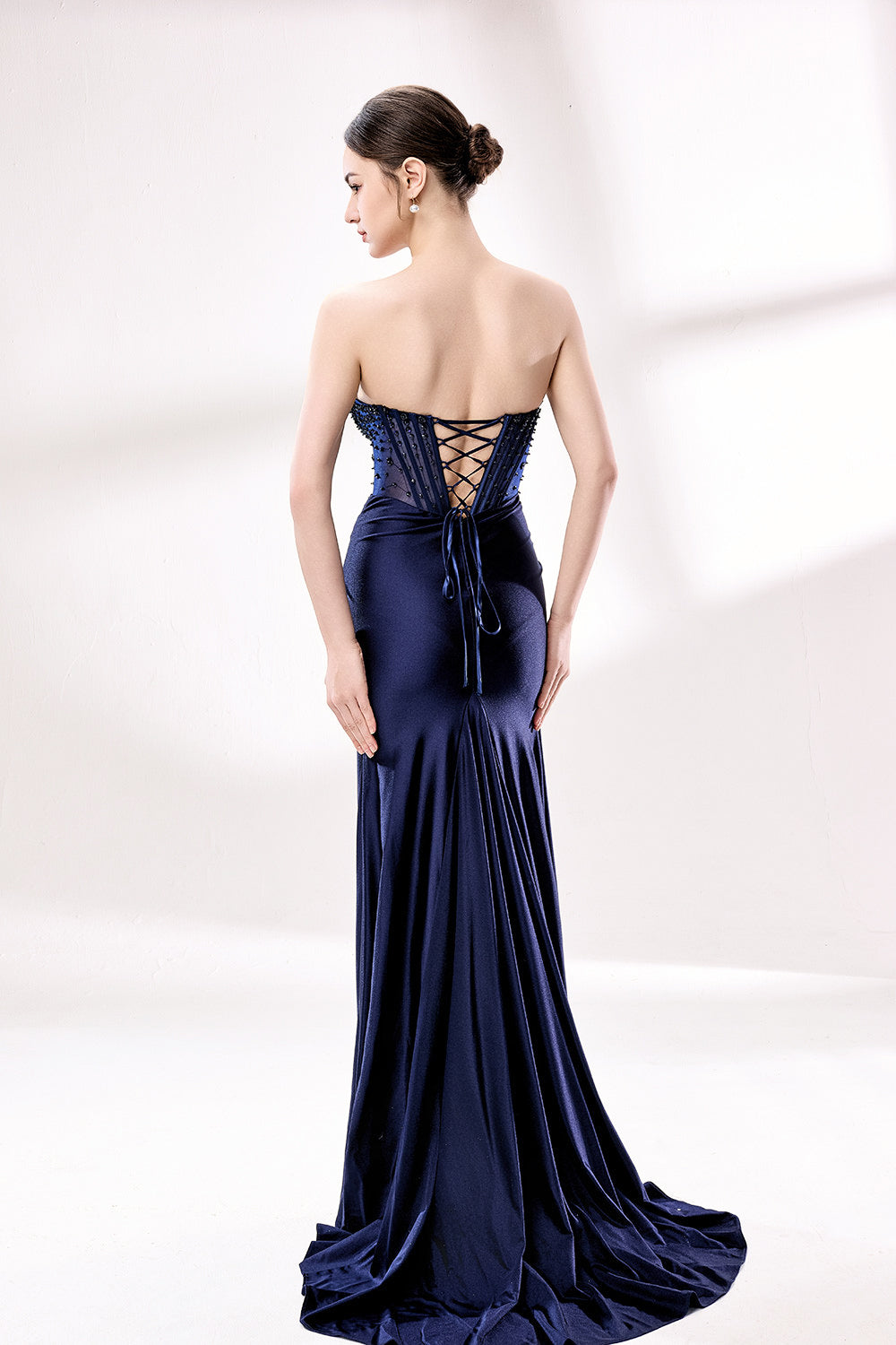 photograph, beautiful young woman, slim figure, long dark hair, fair skin, elegant navy blue strapless gown, fitted bodice with sparkling embellishments, high slit revealing leg, silver high heels, standing pose, white studio background, soft lighting, sophisticated, glamorous, fashion, evening wear, full body shot, elegant fashion photography, modern style, minimalistic setting, stylish, chic.33016-1-02