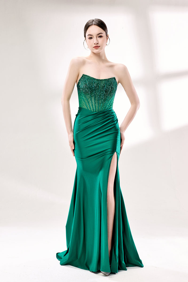 Photograph of a young woman with fair skin and dark hair styled in an elegant updo. She wears a stunning, floor-length, strapless emerald green gown with intricate lace detailing on the bodice and a high slit on the right leg. The dress features a fitted bodice and a flowing, ruched skirt. She accessorizes with large hoop earrings. The background is a soft, white studio setting with diffused lighting, creating a clean, minimalist atmosphere. The woman stands confidently, hands on her hips, exuding grace and