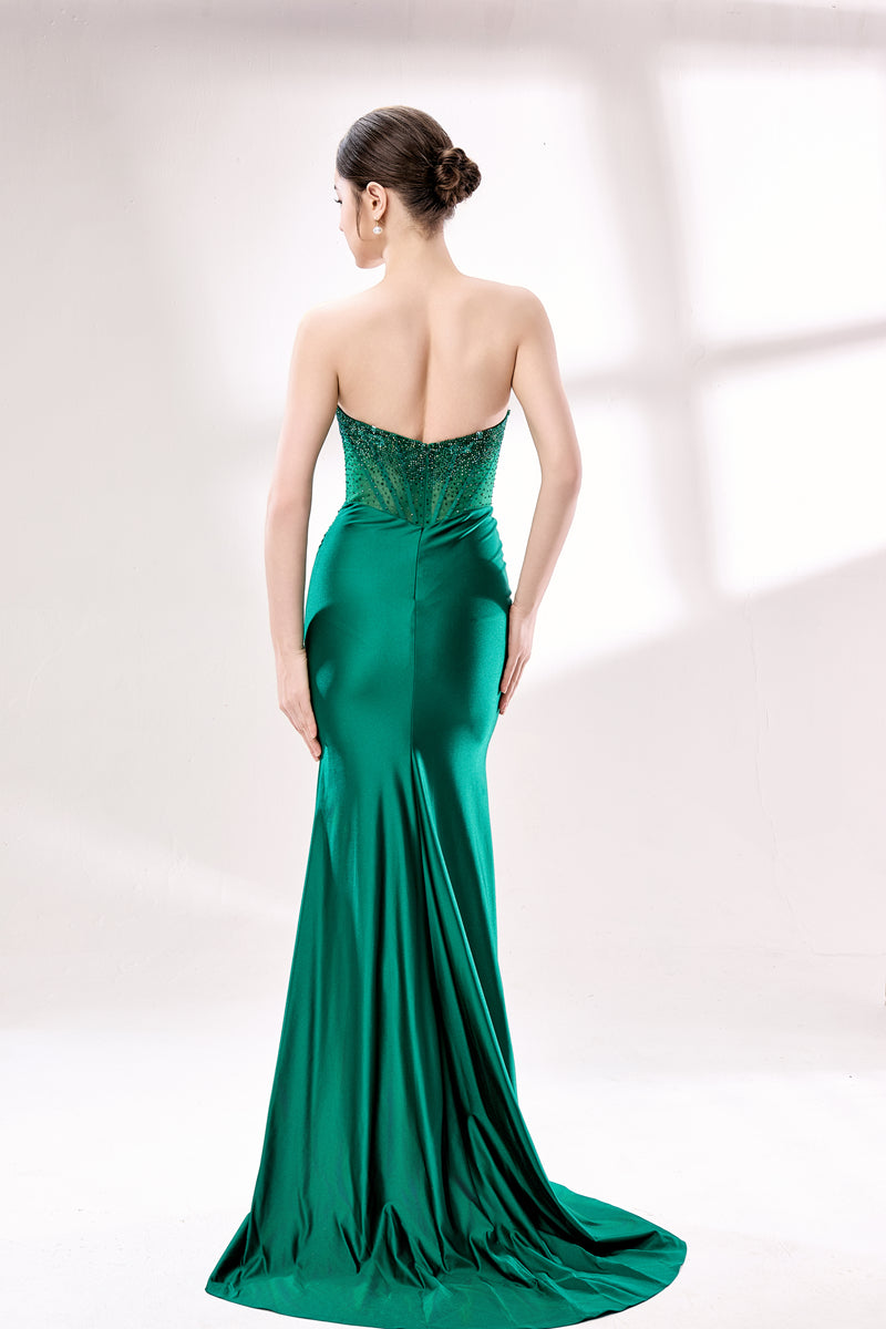 This photograph features a woman with fair skin and dark brown hair styled in a sleek, low bun. She is wearing a strapless, emerald green satin gown with intricate lace detailing at the back, which accentuates her slender figure. The gown has a fitted bodice and a flowing, floor-length skirt. The background is a plain, soft white with diffused lighting, creating a minimalistic and elegant atmosphere. The image is well-lit, highlighting the luxurious texture and color of the dress.