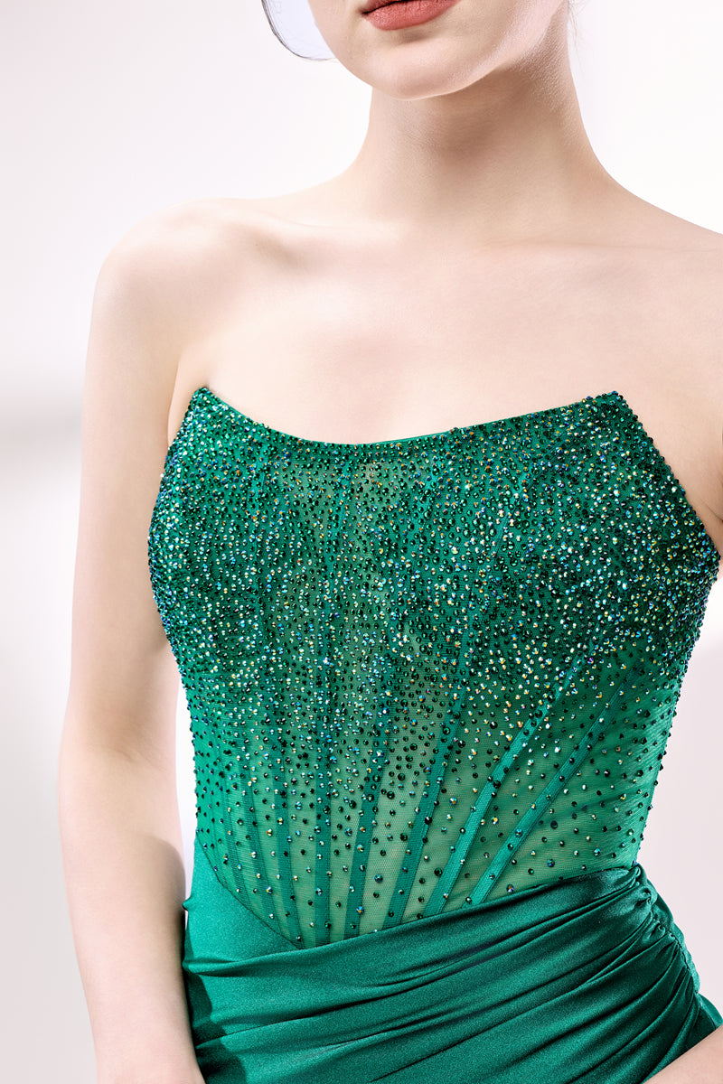 This photograph showcases a close-up of a woman's torso and upper chest. She is wearing a strapless, form-fitting, emerald green dress adorned with intricate beadwork and sequins that shimmer in the light. The dress has a sheer, ruched texture that accentuates her slender figure. Her skin is fair, and her lips are painted a soft pink. The background is a plain, light grey, ensuring that the vibrant green of her dress stands out prominently. The image has a professional, high-fashion feel, with a focus on el