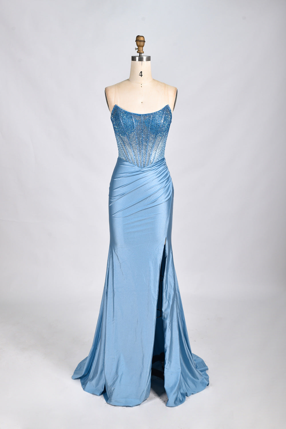 Photograph of a mannequin wearing a strapless, floor-length, royal blue evening gown. The gown features intricate beadwork and embroidery along the bodice, transitioning into a smooth, flowing fabric. The dress has a high slit on the left side, adding a modern touch. The mannequin stands against a plain, light gray background, emphasizing the elegance and sophistication of the gown. The mannequin's head is not visible.