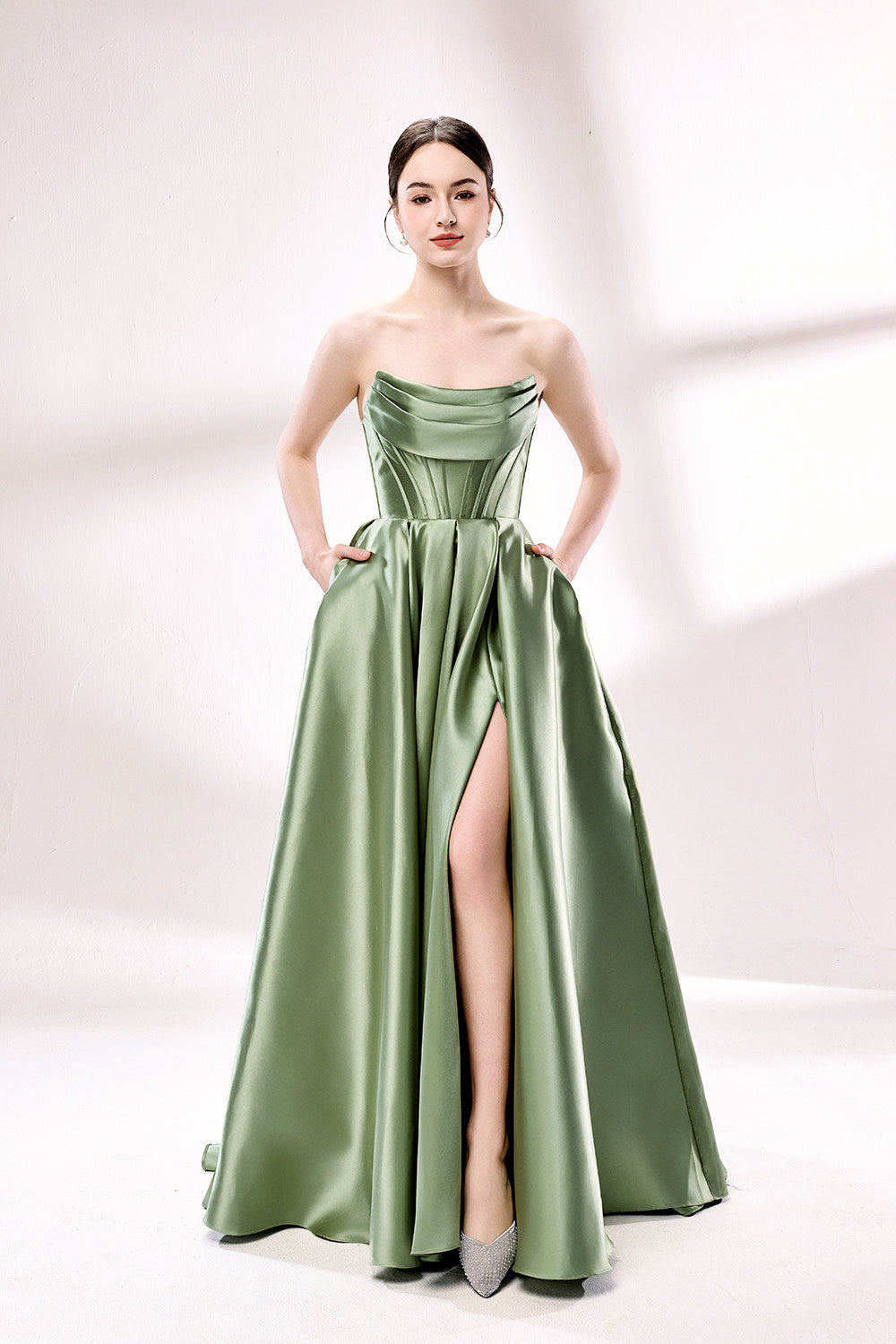 photo of a beautiful young woman standing in front of a white studio background, wearing a strapless, floor-length, olive green satin dress with a high slit on the right leg, hands in pockets, long brunette hair styled in a sleek bun, minimal makeup, elegant and classy look, silver high-heeled shoes, soft lighting, no other objects or people in the frame.33110B-01