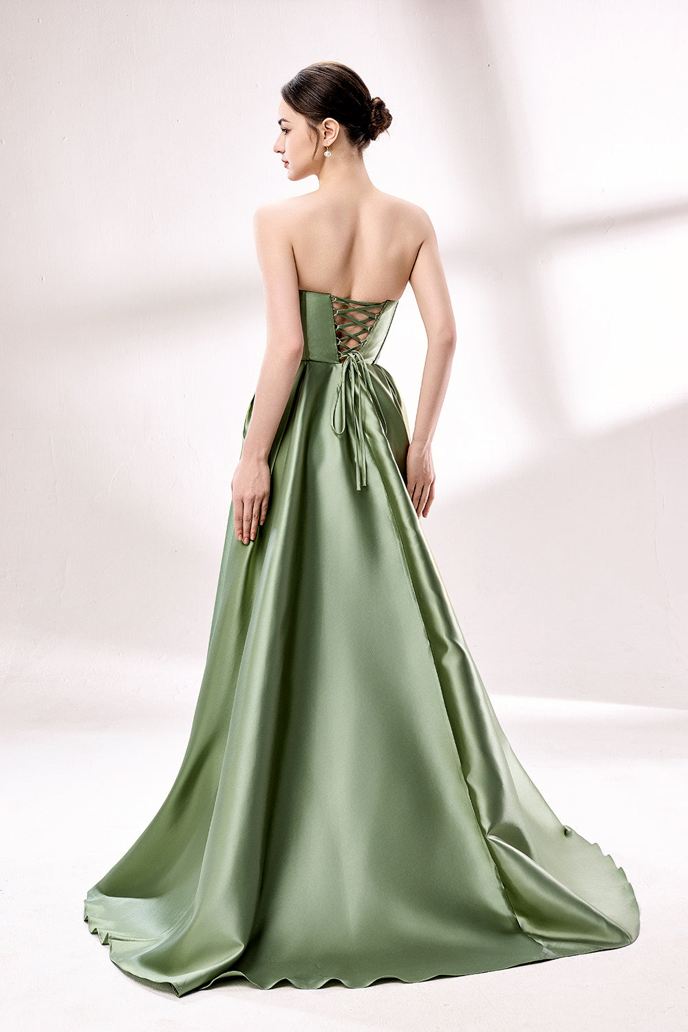 photograph of a woman in a backless, strapless, emerald green satin gown, standing against a white studio background, soft shadows, elegant, classic style, smooth fabric texture, hair in an elegant updo, long flowing gown, no visible accessories, minimalistic setting, studio lighting, elegant and sophisticated, full-length dress, high slit, backless design, full body shot, serene and graceful atmosphere.33110B-02