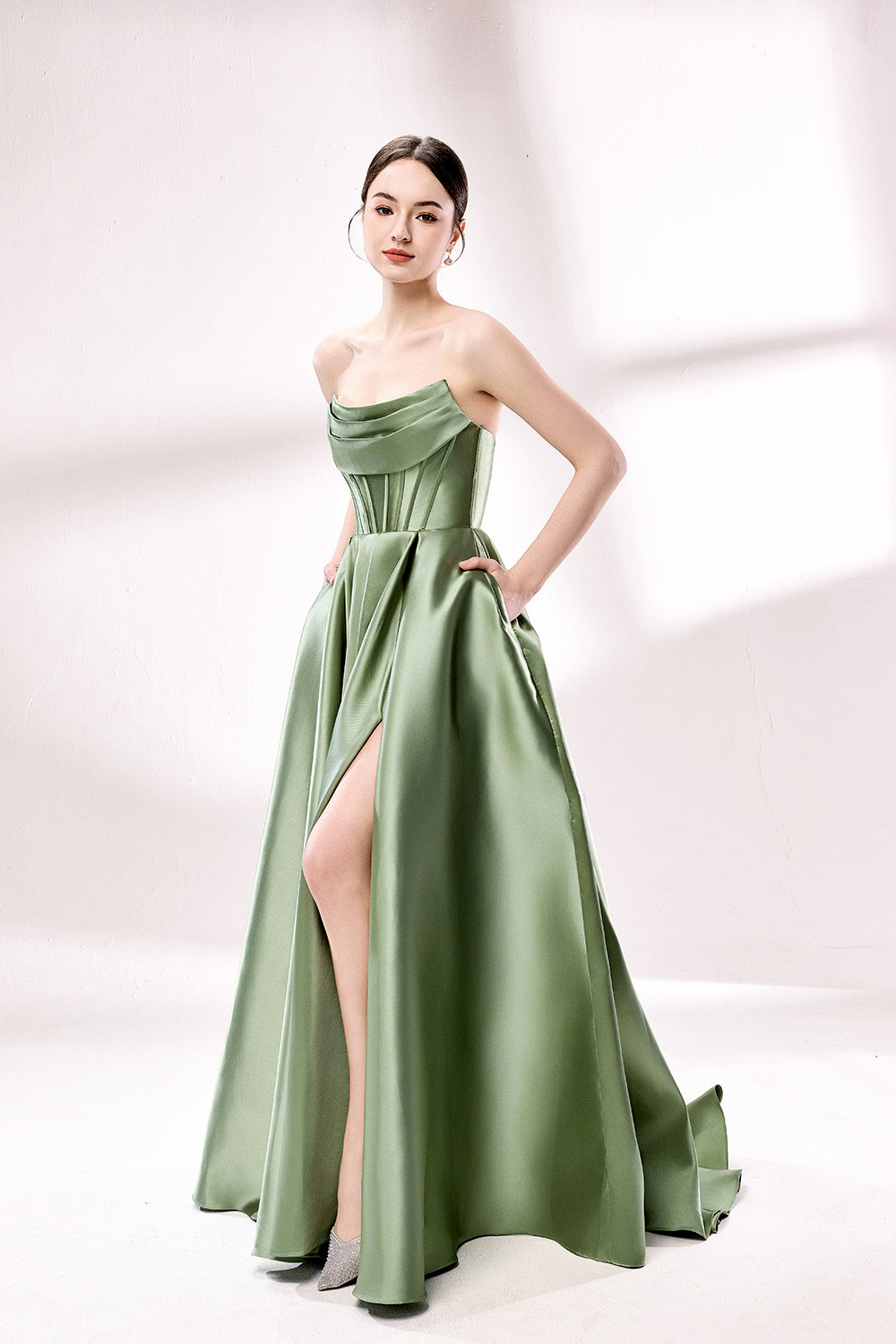 photo of a beautiful young woman standing in front of a white studio background, wearing a strapless, floor-length, olive green satin dress with a high slit on the right leg, hands in pockets, long brunette hair styled in a sleek bun, minimal makeup, elegant and classy look, silver high-heeled shoes, soft lighting, no other objects or people in the frame.33110B-03