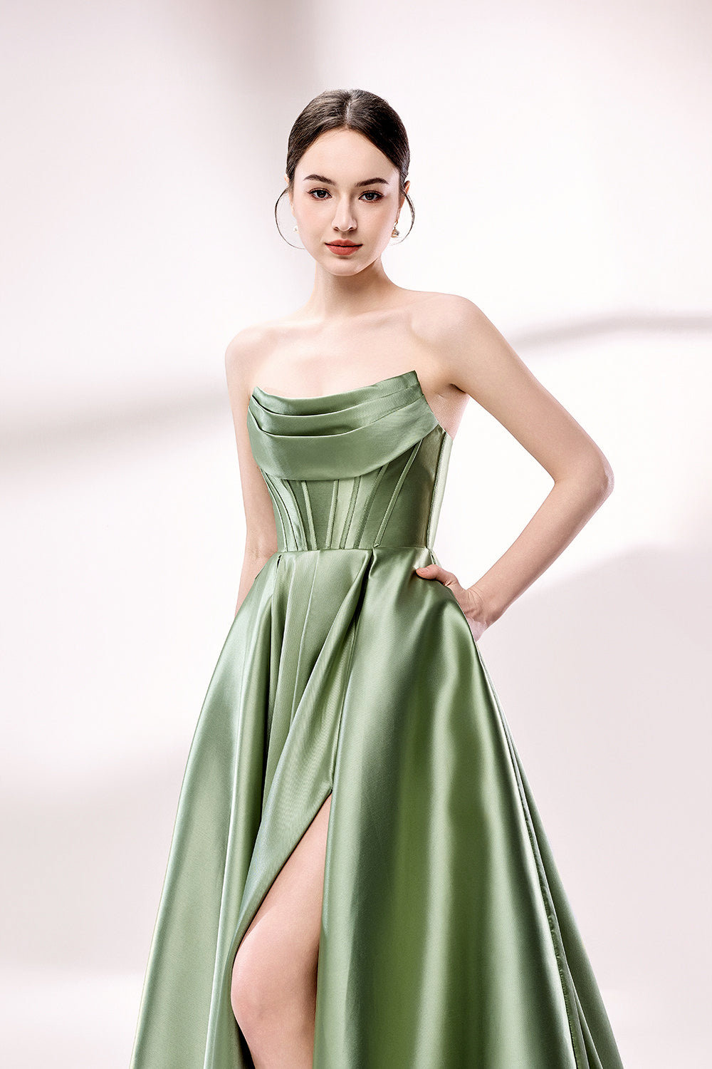 photo of a beautiful young woman standing in front of a white studio background, wearing a strapless, floor-length, olive green satin dress with a high slit on the right leg, hands in pockets, long brunette hair styled in a sleek bun, minimal makeup, elegant and classy look, silver high-heeled shoes, soft lighting, no other objects or people in the frame.33110B-04