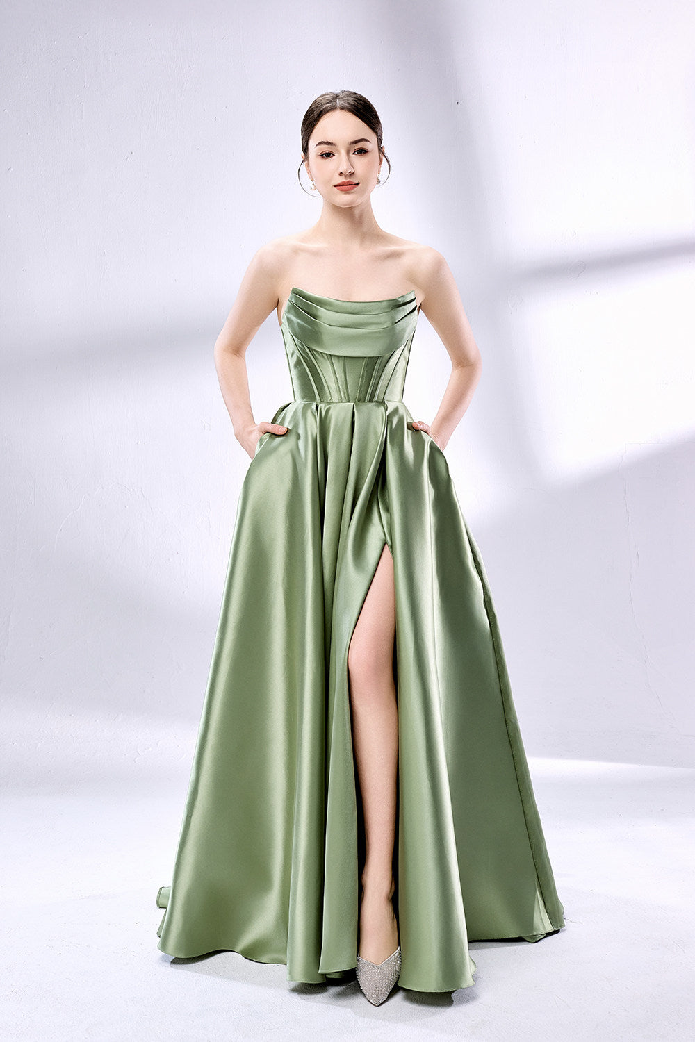 photograph of a beautiful young woman, light skin, brunette, elegant, wearing a strapless, floor-length, emerald green satin gown with a high slit, hands in pockets, standing confidently, soft lighting, white background, shadows on wall, minimalistic setting, fashion shoot, glamour, sophisticated, stylish, modern, chic, elegant dress, high heel shoes, full body shot, studio photography, clean background