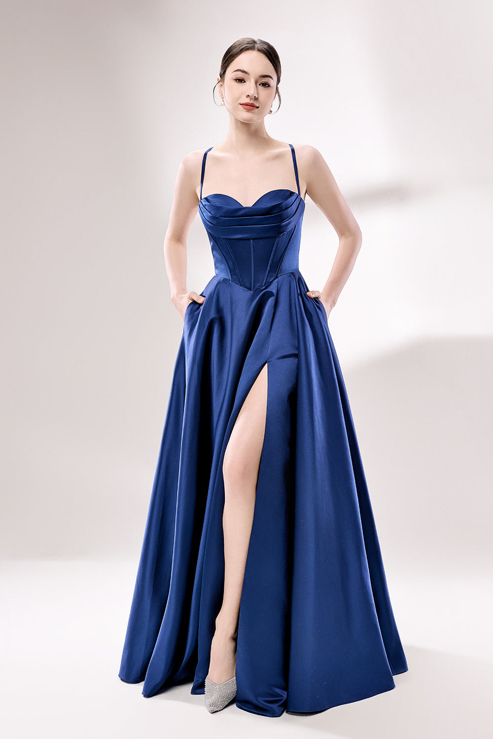 photo of a beautiful young woman, Asian descent, fair skin, long black hair in a low bun, wearing a stunning royal blue satin gown, corset-style top, full skirt, elegant and classy, standing against a plain white background, soft lighting, minimalistic setting, hands behind her back, confident pose, fashion photography, high-quality image, no watermarks.33119-04