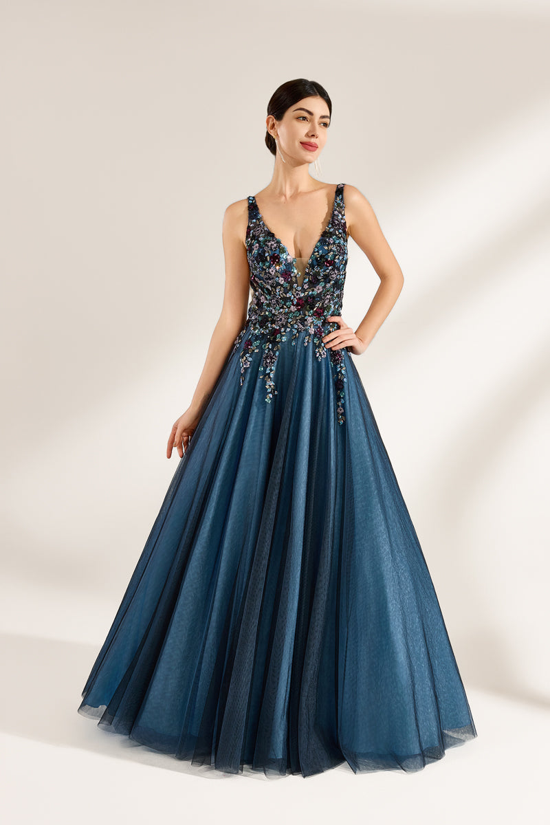 A photograph of a woman in an elegant, floor-length, sleeveless gown. The dress features a deep V-neck adorned with intricate beadwork and sequins in shades of blue, green, and gold. The bodice is fitted, transitioning into a voluminous, blue tulle skirt. Her dark hair is styled in a sleek bun, and she wears gold earrings. The background is a plain, light beige, allowing the dress's details to stand out.