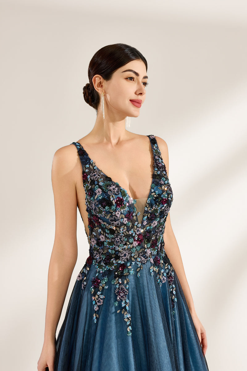 A photograph of a woman in an elegant, floor-length, sleeveless gown. The dress features a deep V-neck adorned with intricate beadwork and sequins in shades of blue, green, and gold. The bodice is fitted, transitioning into a voluminous, blue tulle skirt. Her dark hair is styled in a sleek bun, and she wears gold earrings. The background is a plain, light beige, allowing the dress's details to stand out.