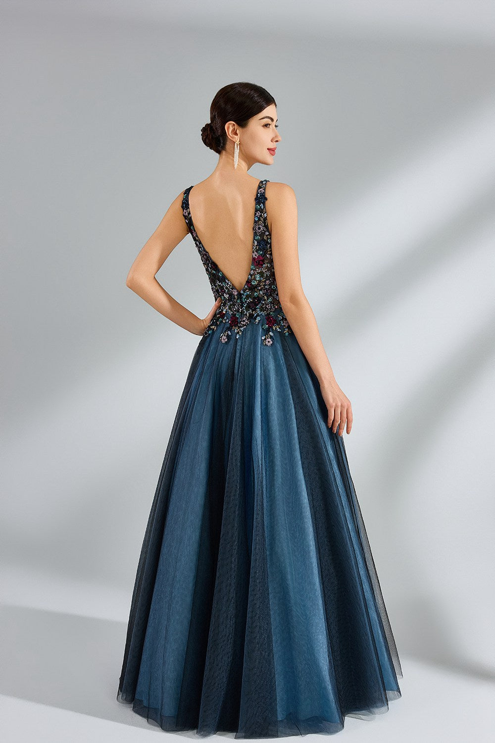Photograph of a woman in an elegant, floor-length, backless gown. The dress features a deep V-back adorned with intricate floral embroidery in shades of red, blue, and green, complementing the blue tulle skirt. Her dark hair is styled in an elegant updo. She stands against a plain, off-white background, with soft, diffused lighting creating subtle shadows. The overall style is sophisticated and glamorous, suitable for formal events.
