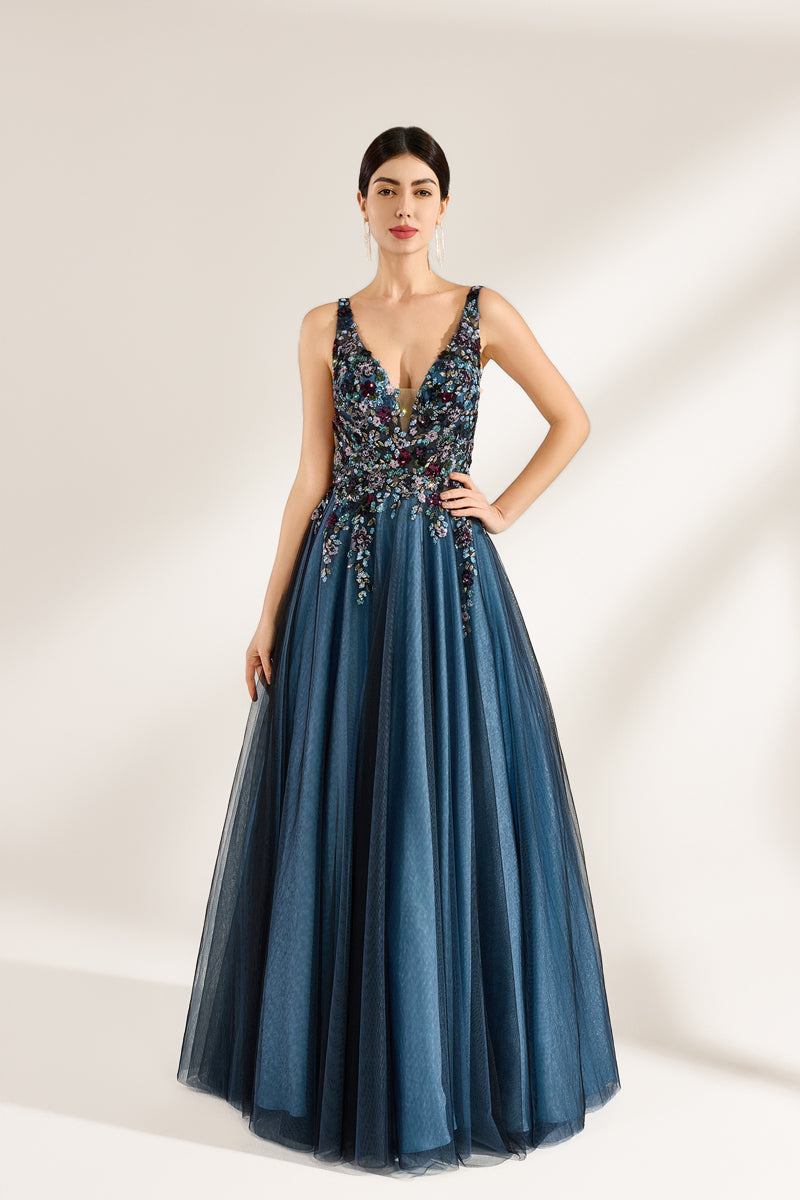 A photograph of a woman in an elegant, floor-length, sleeveless gown. The dress features a deep V-neck adorned with intricate beadwork and sequins in shades of blue, green, and gold. The bodice is fitted, transitioning into a voluminous, blue tulle skirt. Her dark hair is styled in a sleek bun, and she wears gold earrings. The background is a plain, light beige, allowing the dress's details to stand out.