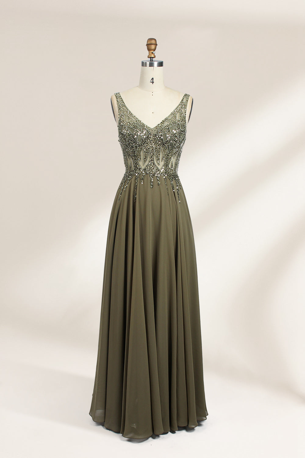 Photograph of an elegant, sleeveless, floor-length gown displayed on a dress form. The gown is a rich olive green with a fitted bodice adorned with intricate, sparkling embellishments. The neckline is a deep V, and the waistline is subtly cinched. The dress flows into a soft, gathered skirt. The background is a plain, light beige, enhancing the gown's luxurious texture and shimmering details.01