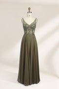 Photograph of an elegant, sleeveless, floor-length gown displayed on a dress form. The gown is a rich olive green with a fitted bodice adorned with intricate, sparkling embellishments. The neckline is a deep V, and the waistline is subtly cinched. The dress flows into a soft, gathered skirt. The background is a plain, light beige, enhancing the gown's luxurious texture and shimmering details.01