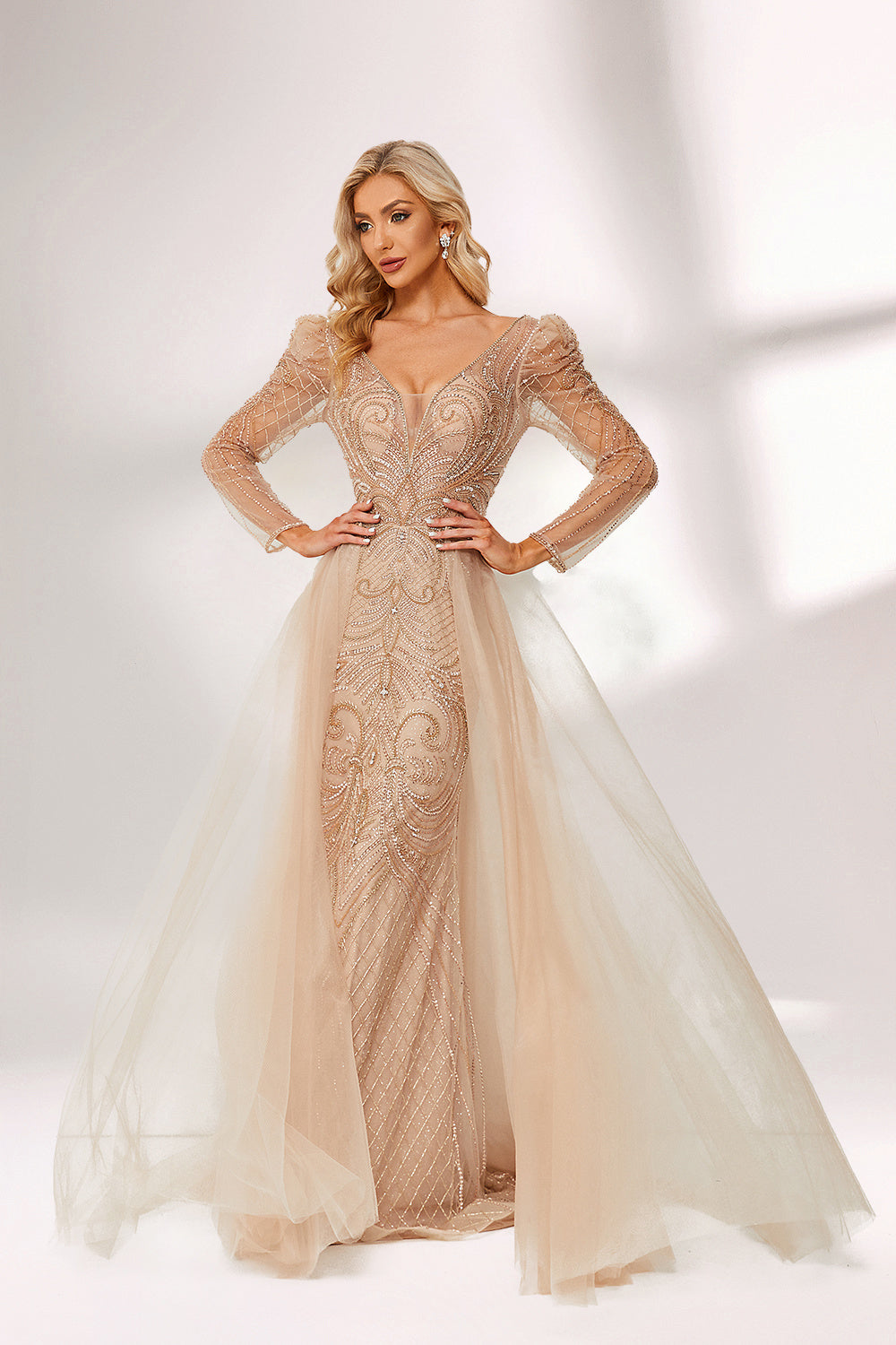 A woman stands against a light background, wearing a champagne - colored mermaid - style gown. It features a deep V - neckline, intricate beadwork throughout, and long sheer sleeves. The elegant design creates a glamorous and sophisticated look, perfect for formal events like galas or weddings.5