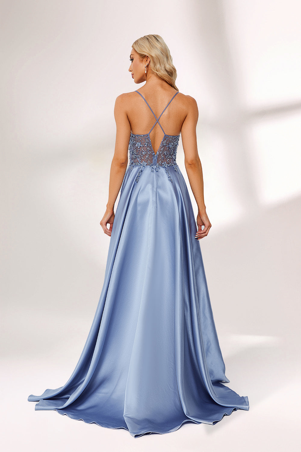Photograph of a tall, slender blonde woman with fair skin, wearing a floor-length, light blue satin gown with a plunging V-neckline and intricate lace detailing. She has a high slit on one side, revealing a silver high-heeled shoe. The background is a simple, white studio setting with soft lighting.06