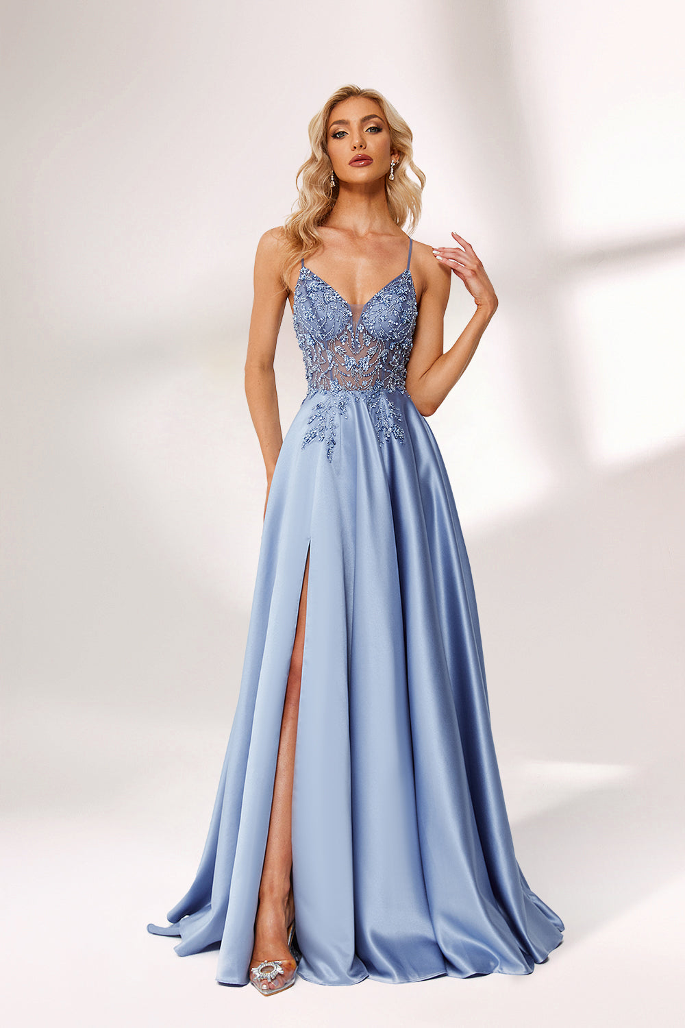 Photograph of a tall, slender blonde woman with fair skin, wearing a floor-length, light blue satin gown with a plunging V-neckline and intricate lace detailing. She has a high slit on one side, revealing a silver high-heeled shoe. The background is a simple, white studio setting with soft lighting.01
