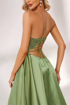 In soft lighting, a woman stuns in a green evening gown. It has spaghetti straps, a lace bodice, and a satin skirt with a slit, creating an elegant and alluring look for formal events.4