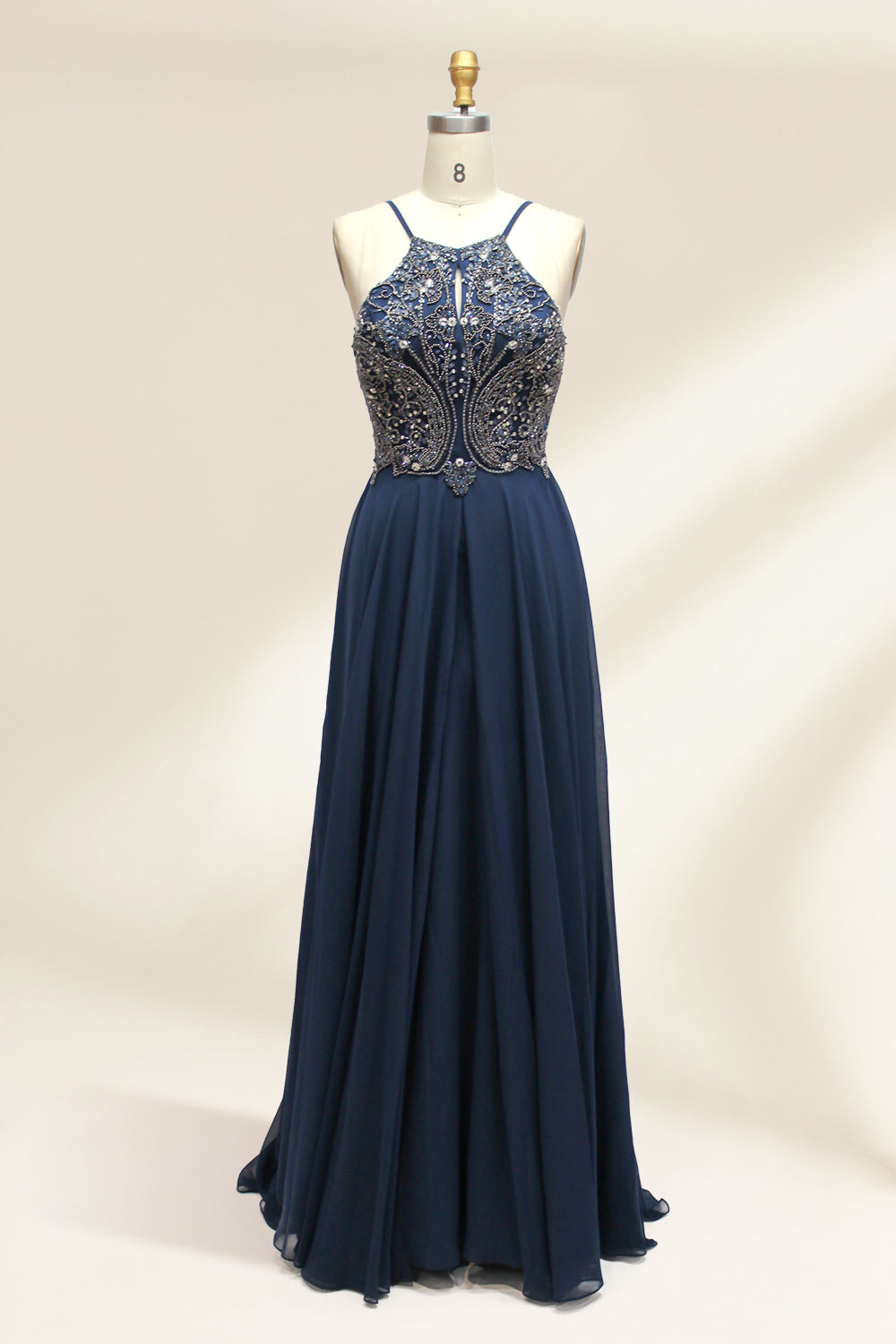 Photograph of a sleeveless, floor-length navy blue gown on a dress form. The gown features intricate beadwork and sequin embroidery on the bodice, with a sheer, flowing skirt. The dress form is positioned against a plain white background, highlighting the elegant and luxurious design of the dress.