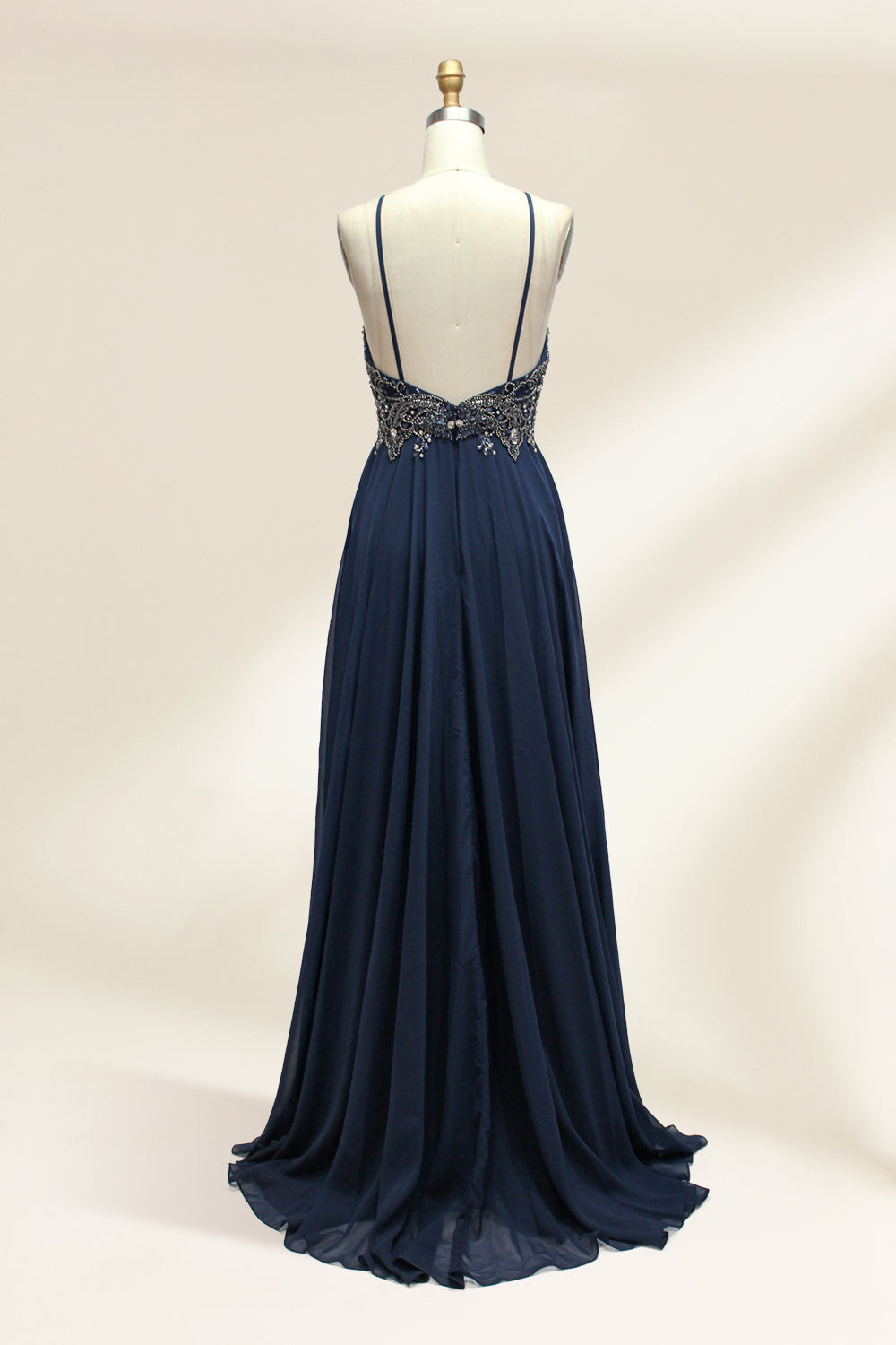 Photograph of a navy blue, floor-length, sleeveless gown on a dress form. The dress features intricate beadwork and sequin embellishments on the bodice, with a flowing, sheer skirt. The form is placed against a plain, light beige background, highlighting the dress's elegant, sophisticated design.9107-02