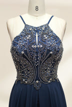 Photograph of a navy blue, floor-length, sleeveless gown on a dress form. The dress features intricate beadwork and sequin embellishments on the bodice, with a flowing, sheer skirt. The form is placed against a plain, light beige background, highlighting the dress's elegant, sophisticated design.9107-03