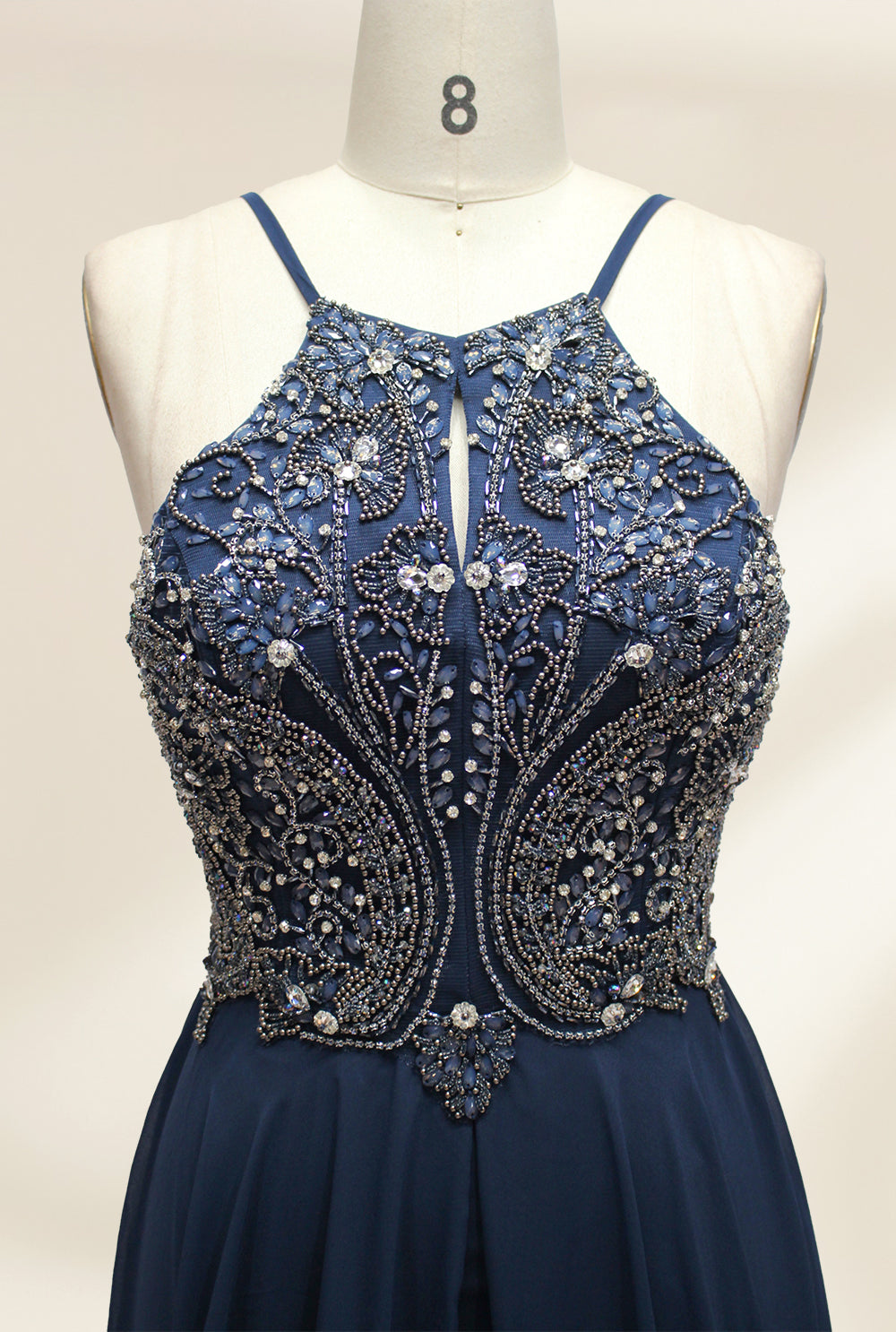 Photograph of a navy blue, floor-length, sleeveless gown on a dress form. The dress features intricate beadwork and sequin embellishments on the bodice, with a flowing, sheer skirt. The form is placed against a plain, light beige background, highlighting the dress's elegant, sophisticated design.9107-03