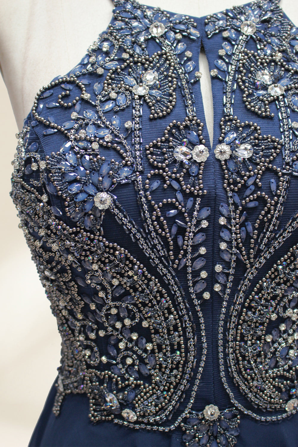Photograph of a navy blue, floor-length, sleeveless gown on a dress form. The dress features intricate beadwork and sequin embellishments on the bodice, with a flowing, sheer skirt. The form is placed against a plain, light beige background, highlighting the dress's elegant, sophisticated design.9107-04