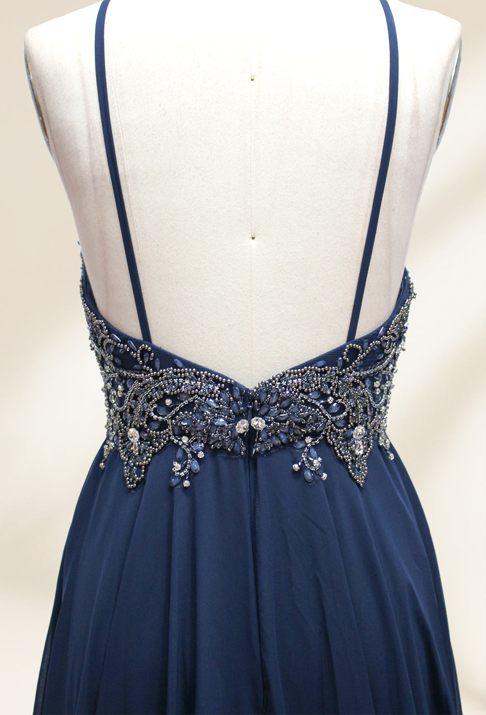 Photograph of a navy blue, floor-length, sleeveless gown on a dress form. The dress features intricate beadwork and sequin embellishments on the bodice, with a flowing, sheer skirt. The form is placed against a plain, light beige background, highlighting the dress's elegant, sophisticated design.9107-05