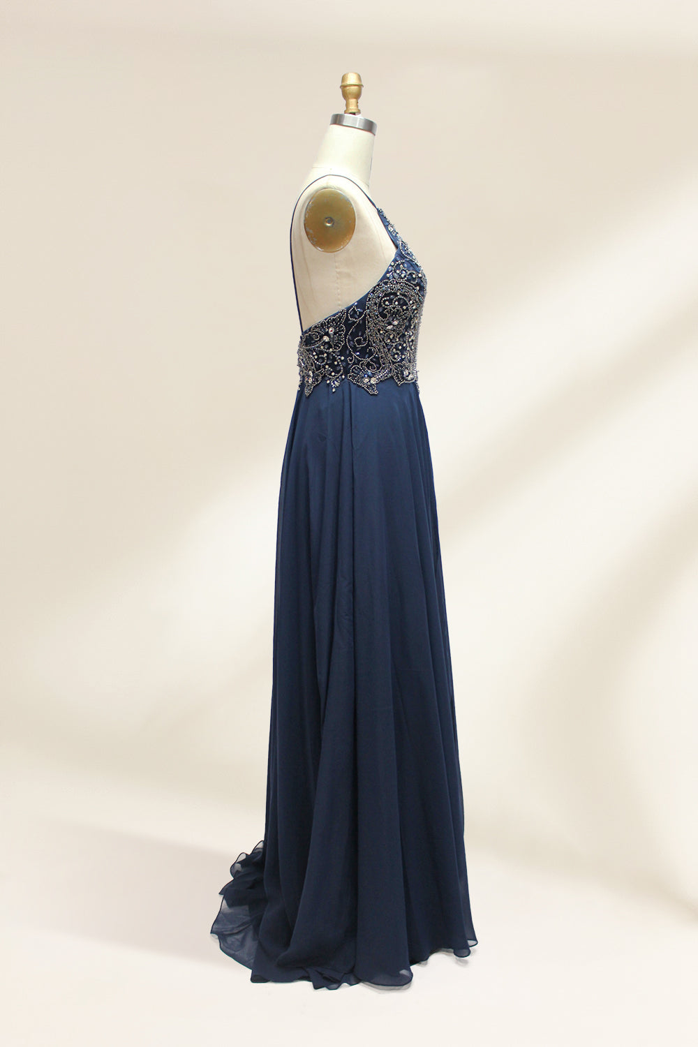 Photograph of a navy blue, floor-length, sleeveless gown on a dress form. The dress features intricate beadwork and sequin embellishments on the bodice, with a flowing, sheer skirt. The form is placed against a plain, light beige background, highlighting the dress's elegant, sophisticated design.9107-06