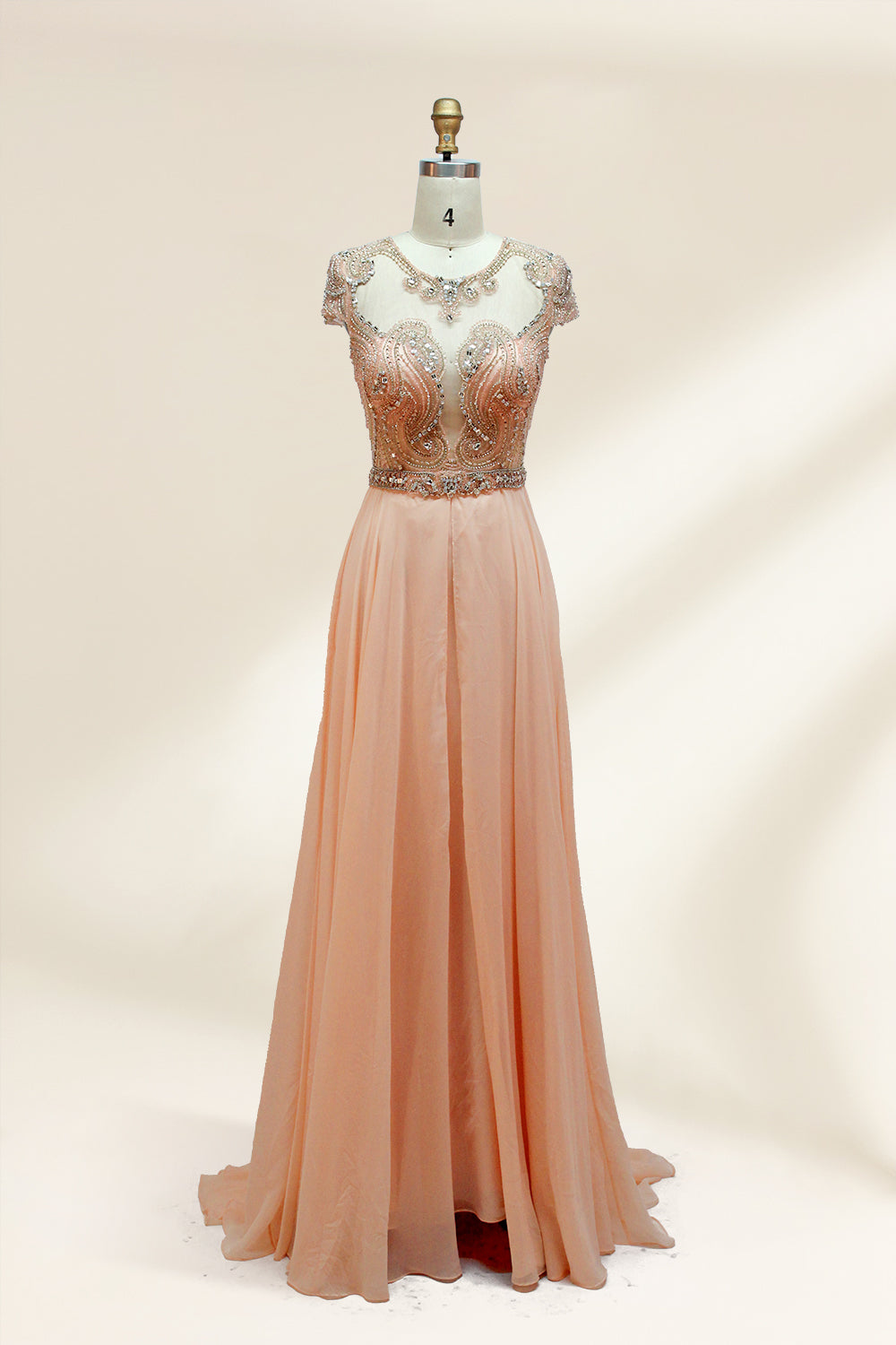 Photograph of an elegant, floor-length, peach-colored evening gown on a mannequin. The gown features intricate, sparkling beadwork on the bodice and cap sleeves, and a flowing, sheer skirt. The mannequin has a light beige background, with soft shadows highlighting the gown's details. The dress exudes luxury and sophistication.9132.1