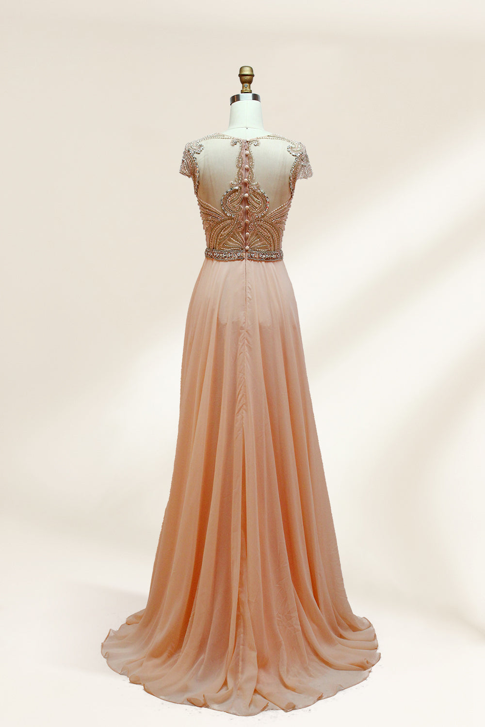 Photograph of an elegant, floor-length, peach-colored evening gown on a mannequin. The gown features intricate, sparkling beadwork on the bodice and cap sleeves, and a flowing, sheer skirt. The mannequin has a light beige background, with soft shadows highlighting the gown's details. The dress exudes luxury and sophistication.9132.2