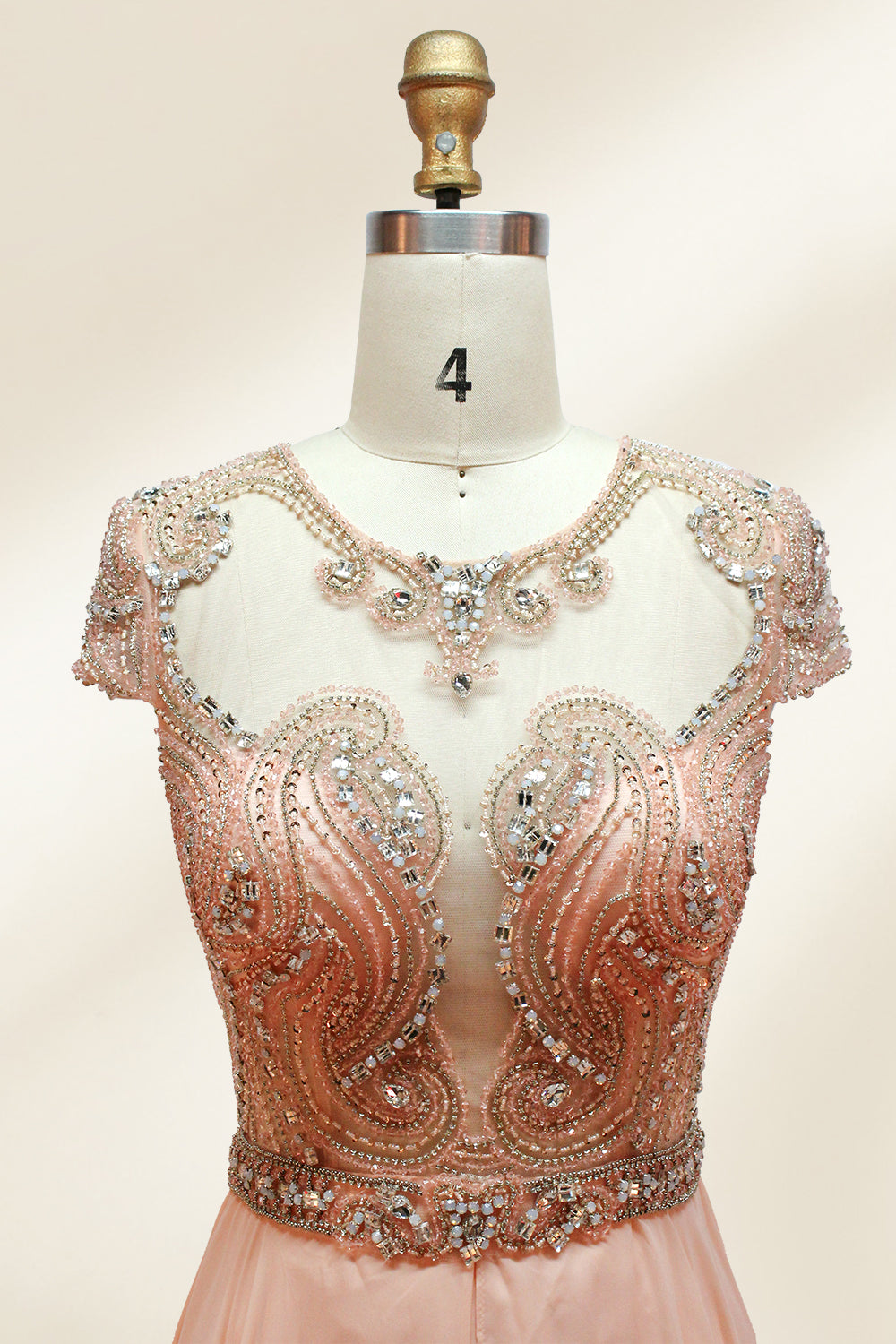 Photograph of an elegant, floor-length, peach-colored evening gown on a mannequin. The gown features intricate, sparkling beadwork on the bodice and cap sleeves, and a flowing, sheer skirt. The mannequin has a light beige background, with soft shadows highlighting the gown's details. The dress exudes luxury and sophistication.9132.3