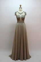 Photograph of an elegant, floor-length, peach-colored evening gown on a mannequin. The gown features intricate, sparkling beadwork on the bodice and cap sleeves, and a flowing, sheer skirt. The mannequin has a light beige background, with soft shadows highlighting the gown's details. The dress exudes luxury and sophistication.9132.7