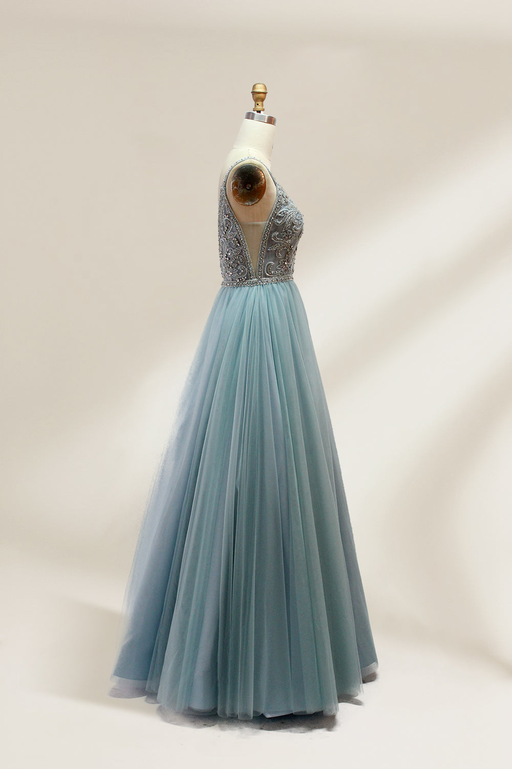 A photograph of a mannequin showcasing a floor-length, light blue tulle gown with intricate silver embroidery on the bodice. The gown has a fitted, sleeveless top and a voluminous skirt. The background is a plain, soft beige, highlighting the gown's elegant and sophisticated design.9133.3