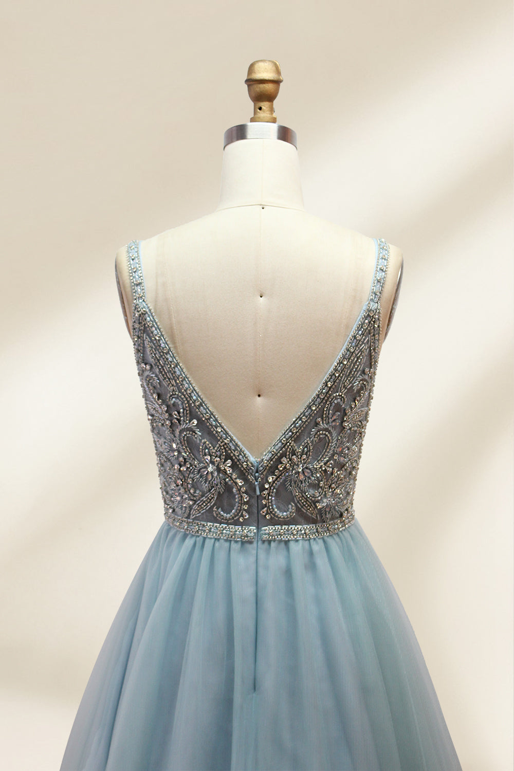 A photograph of a mannequin showcasing a floor-length, light blue tulle gown with intricate silver embroidery on the bodice. The gown has a fitted, sleeveless top and a voluminous skirt. The background is a plain, soft beige, highlighting the gown's elegant and sophisticated design.