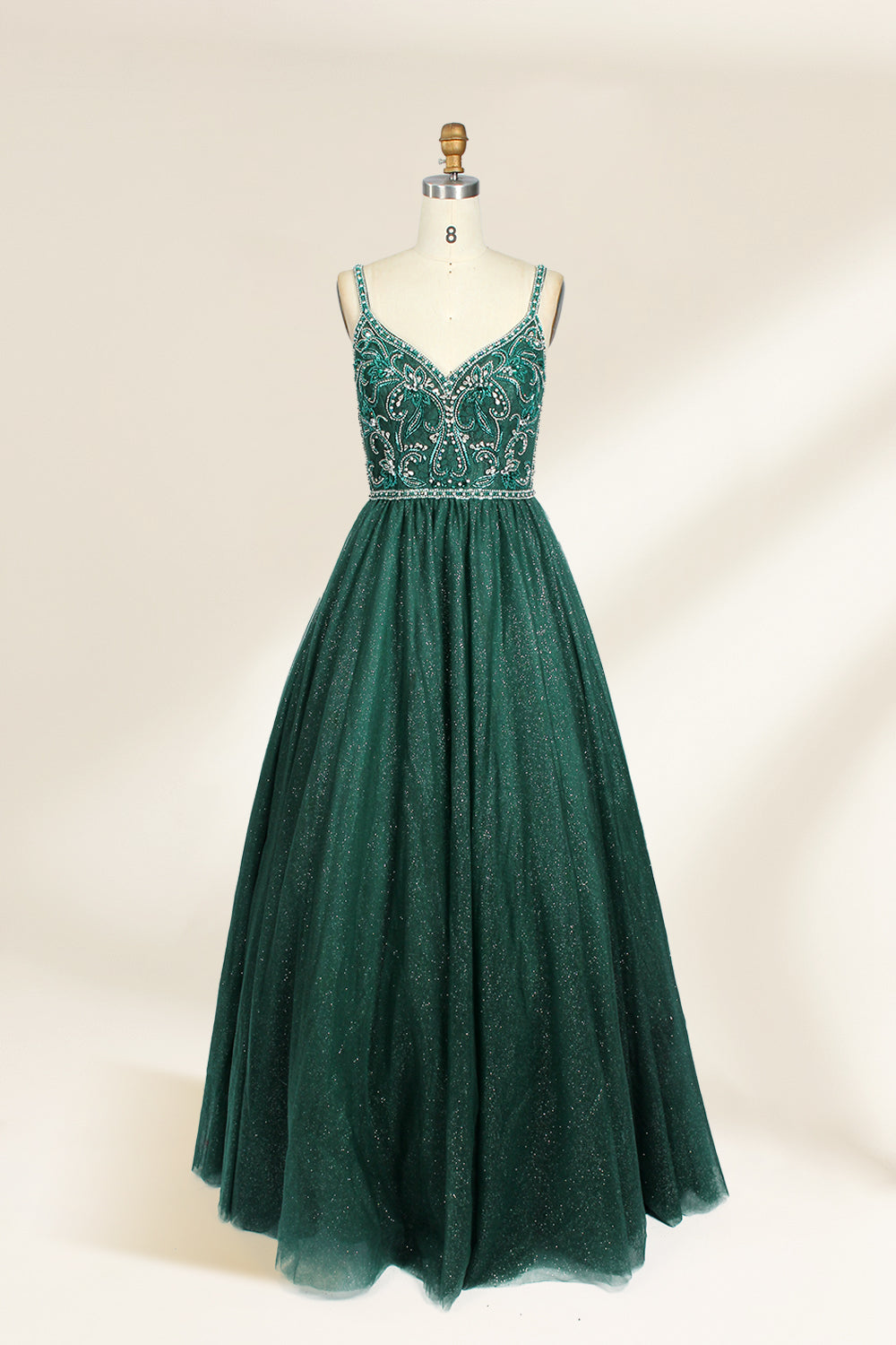 A photograph of a mannequin showcasing a floor-length, sleeveless, dark green evening gown. The gown features a V-neckline embellished with intricate beadwork and a fitted bodice that transitions into a voluminous tulle skirt. The background is plain and off-white, emphasizing the gown's elegant design.9133b.1