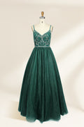 A photograph of a mannequin showcasing a floor-length, sleeveless, dark green evening gown. The gown features a V-neckline embellished with intricate beadwork and a fitted bodice that transitions into a voluminous tulle skirt. The background is plain and off-white, emphasizing the gown's elegant design.9133b.1