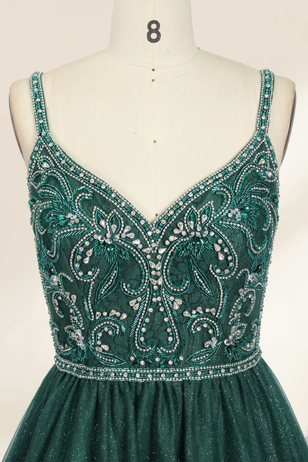 A photograph of a mannequin showcasing a floor-length, sleeveless, dark green evening gown. The gown features a V-neckline embellished with intricate beadwork and a fitted bodice that transitions into a voluminous tulle skirt. The background is plain and off-white, emphasizing the gown's elegant design.9133b.2